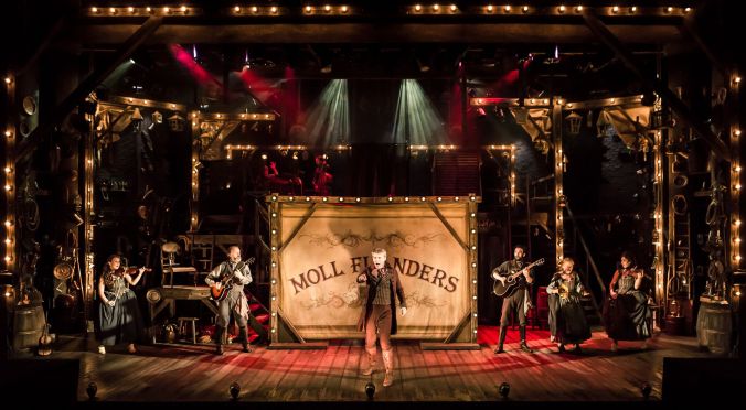 Photo shows full cast of stage, with the character 'Daniel DeFoe' setting the scene. The light is a mixture of reds and oranges. Daniel is standing centre stage with the title 'Moll Flanders' behind him on a canvas.
