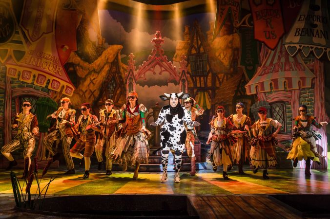 Photo shows a villiage scene with numerous villagers dancing in the background whilst 'Day-Z' the cow sings a musical number