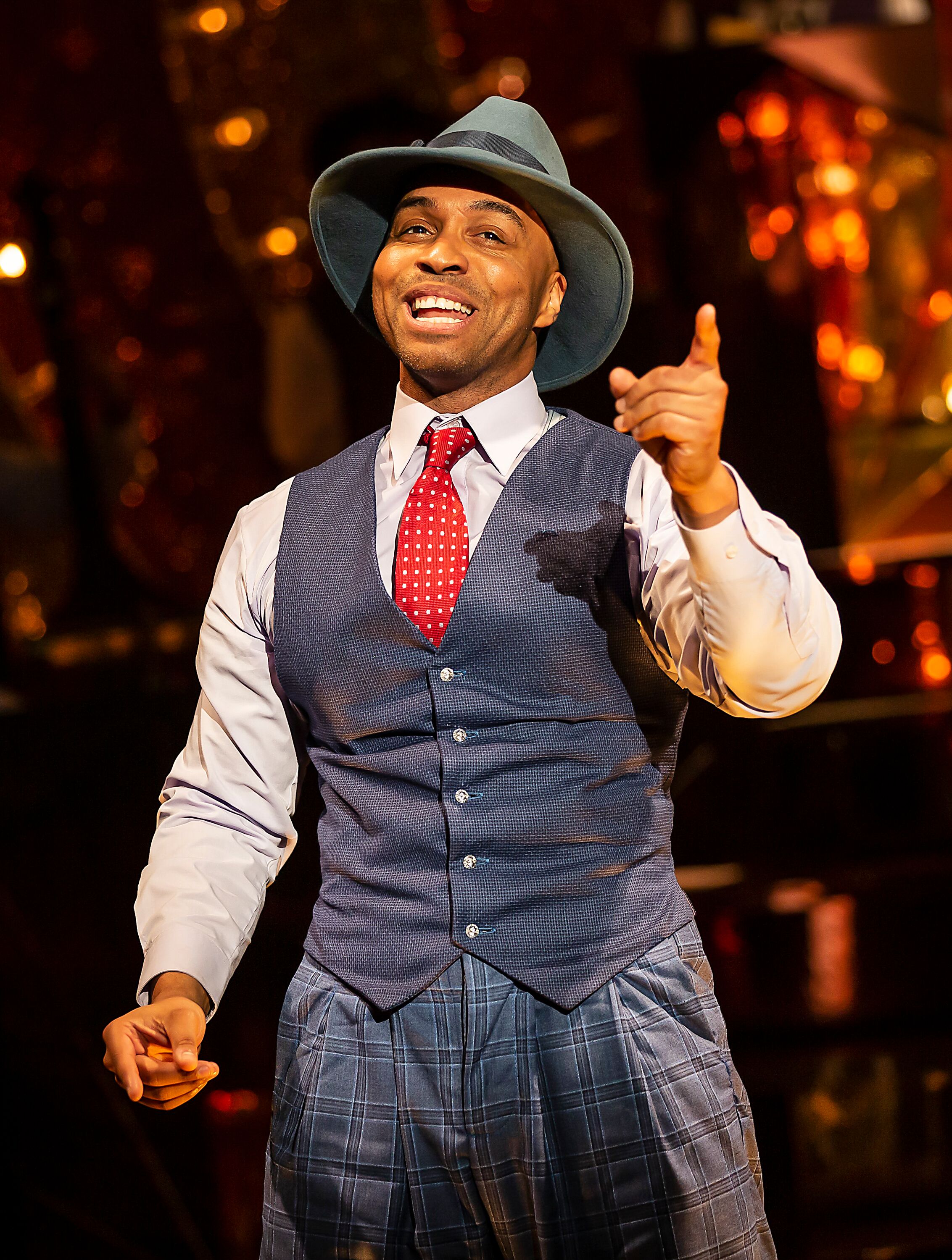 photo shows one of the male singers Wayne Robinson singing to the audience. He is wear a blue wastecoat with blue suit trousers, plain blue shirt, a red white dotted tie and a blue Fedora like hat.
