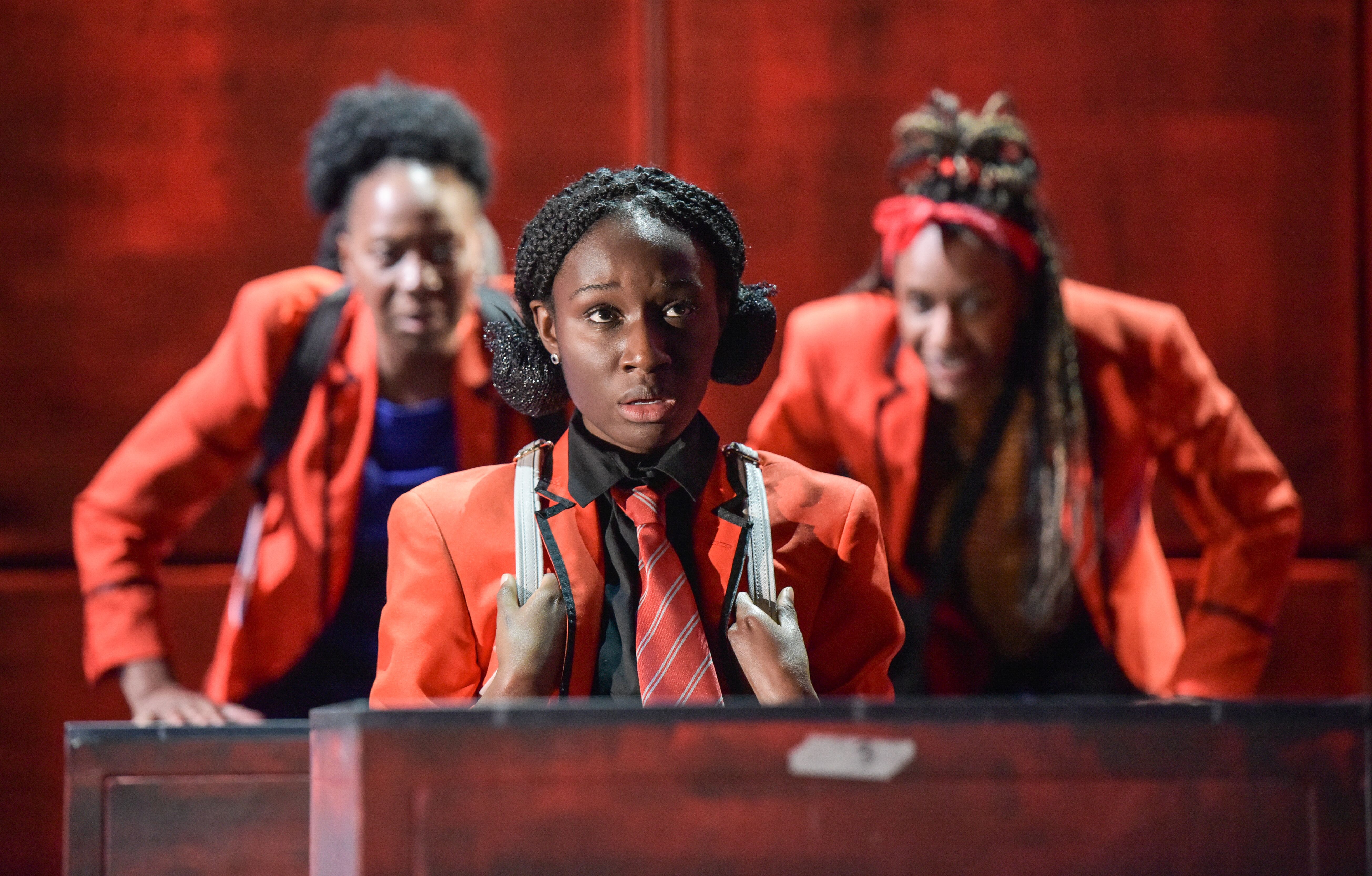 Photo shows Sephy’ (Heather Agyepong) wearing a red school uniform looking into the distance with two other students behind her poking fun of her.