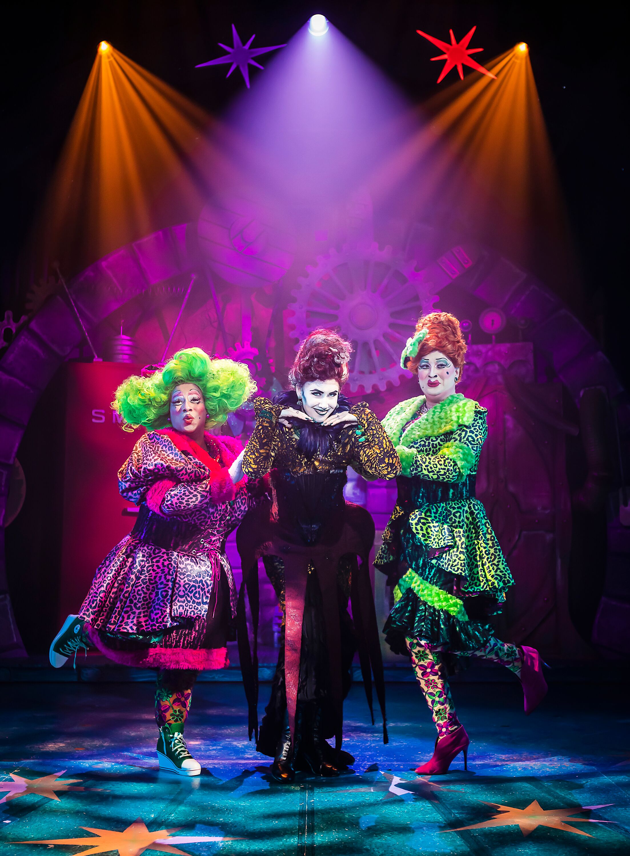 Image shows both the Ugly Step Sister one, in a lime green dress the other in a neon pink dress posing with thier step mother who is wear all black looking into the audience, ornage and pink lights around them. Photography By Pamela Raith