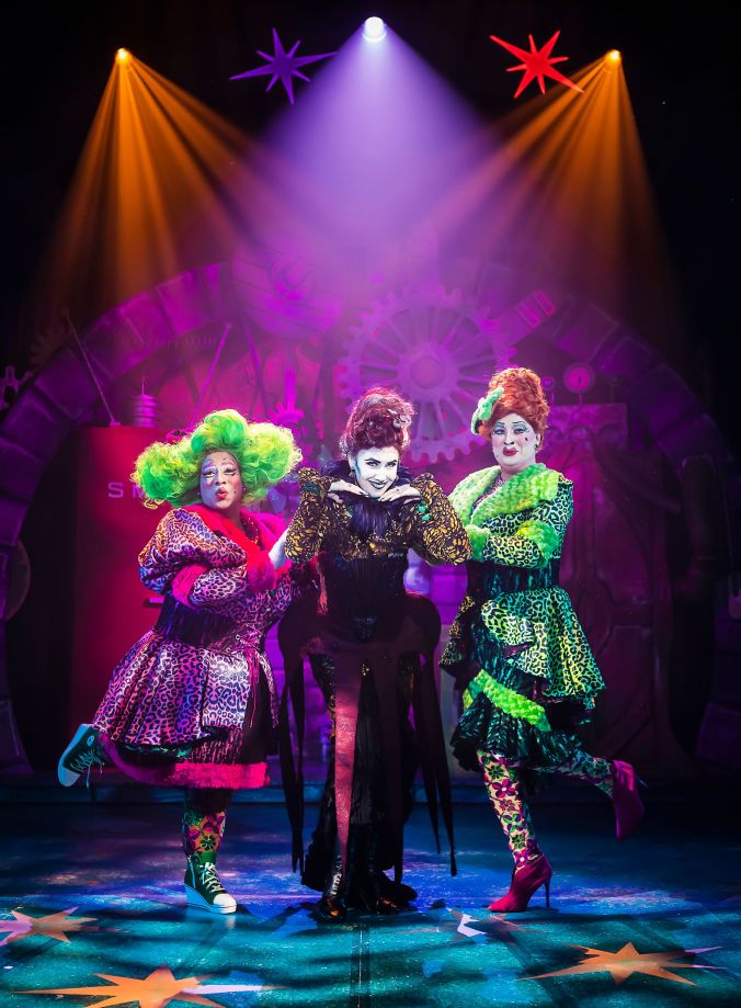 Image shows both the Ugly Step Sister one, in a lime green dress the other in a neon pink dress posing with thier step mother who is wear all black looking into the audience, ornage and pink lights around them. Photography By Pamela Raith