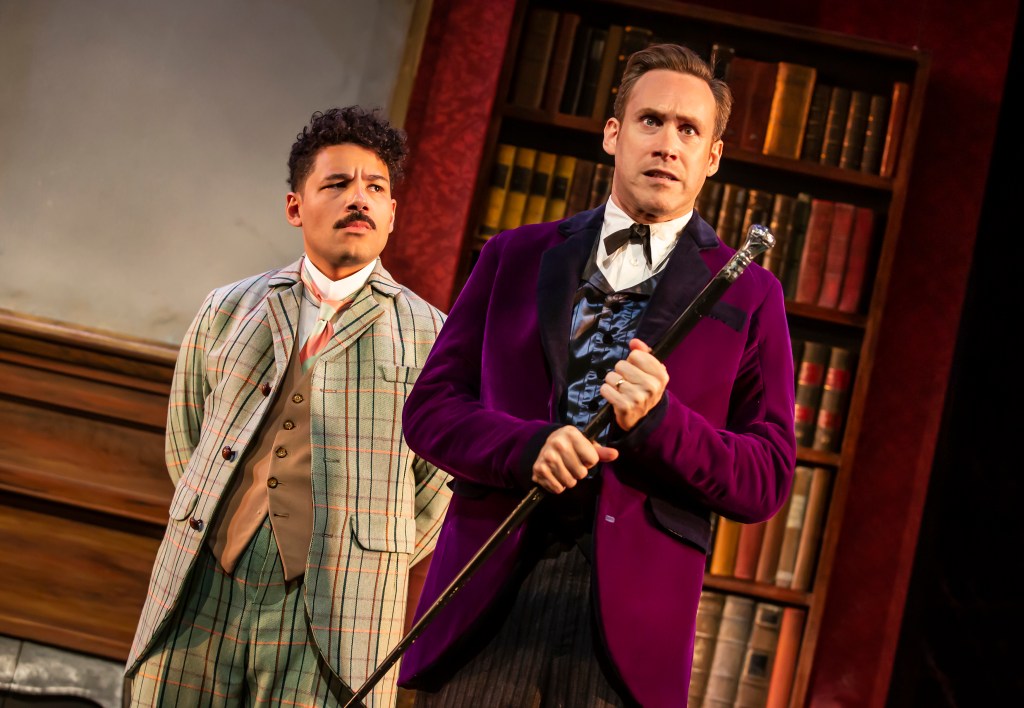 Imagine of Holmes in a purple blazer holding a black and silver cane, looking concerned, with Waston just behind him looking at Holmes expectantly.