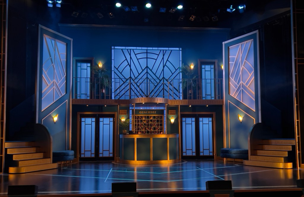 A hotel lobby, with an upper balcony. Underneath, is a reception desk with sets of keys behind it. On both sides of the desk are two big sets of doors. Near the front of the stage are two small staircases, going on and off stage, all of the set. is coloured in gold and blue.