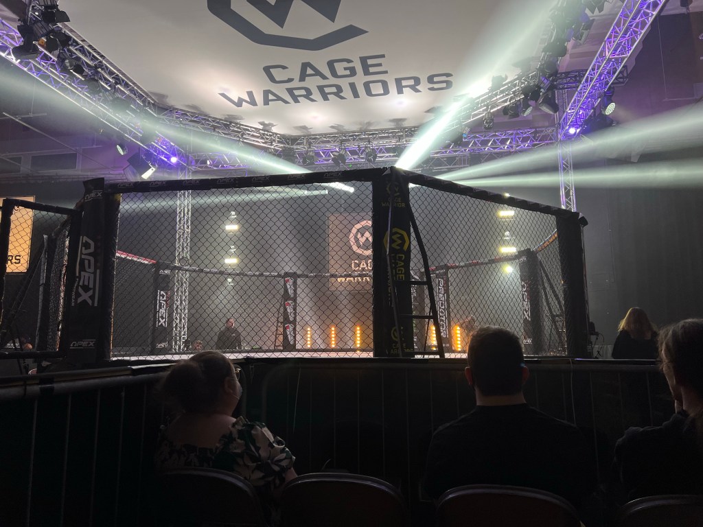 photo shows a photo of the cage warriors fighting game, with white lights flashing around. Gage warriors branding is all around the sides and above the cage with screens all around the room.