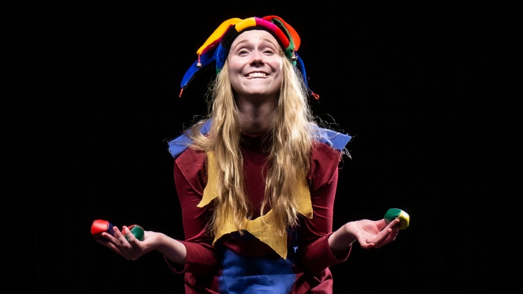 Bard (Megan Risley) is wearing a very colourful court jester outlet whilst smiling juggling some colourful juggling balls.