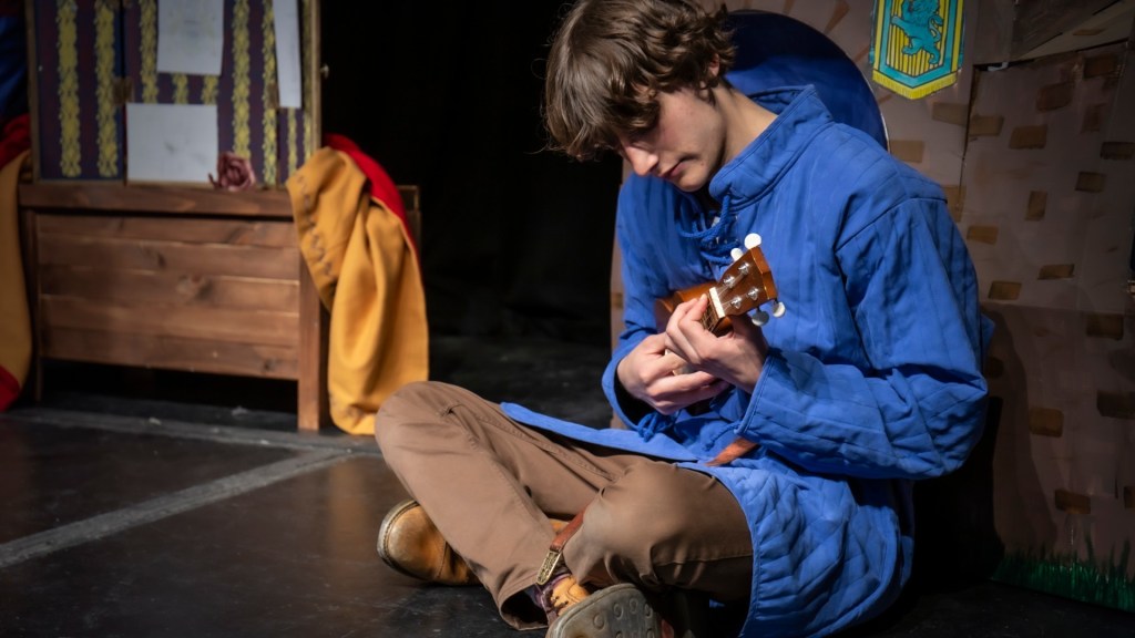 James (Tommy Campe) is sitting cross legged on the floor wearing a blue medieval garment, playing the ukulele.