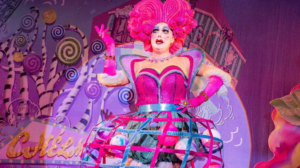 Betty Bon Bon (panto dame) is speaking to the audience wearing a bright pink dress with wig and same gloves the same colour with a dress of a net and ball balls 