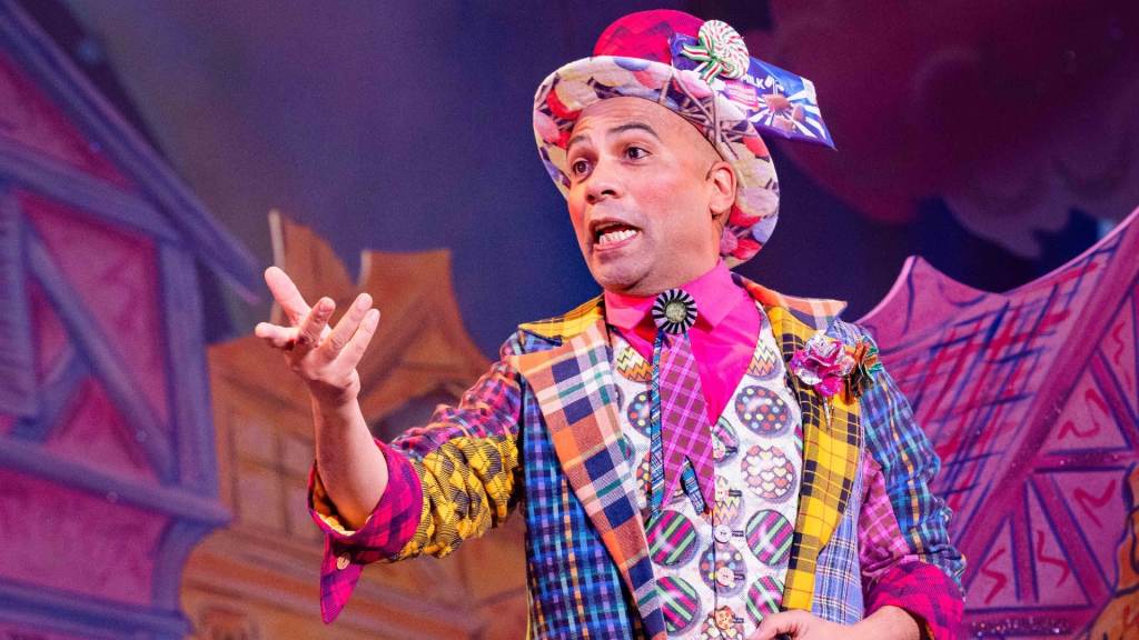 Dale Superville is wearing a multicoloured suit like costume with a multicoloured hat point and talking to another character on stage.