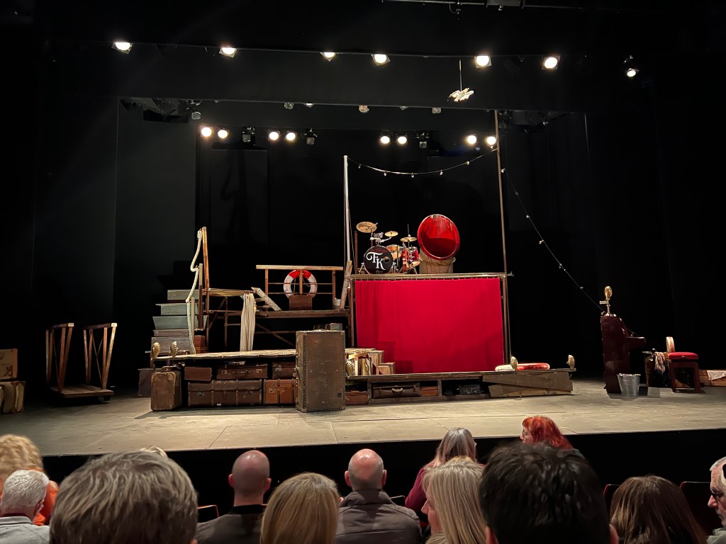 Photo shows a photo of the set before the show. A makeshift boat made of wood planks, with two stories, with the stairs on on the left hand side.