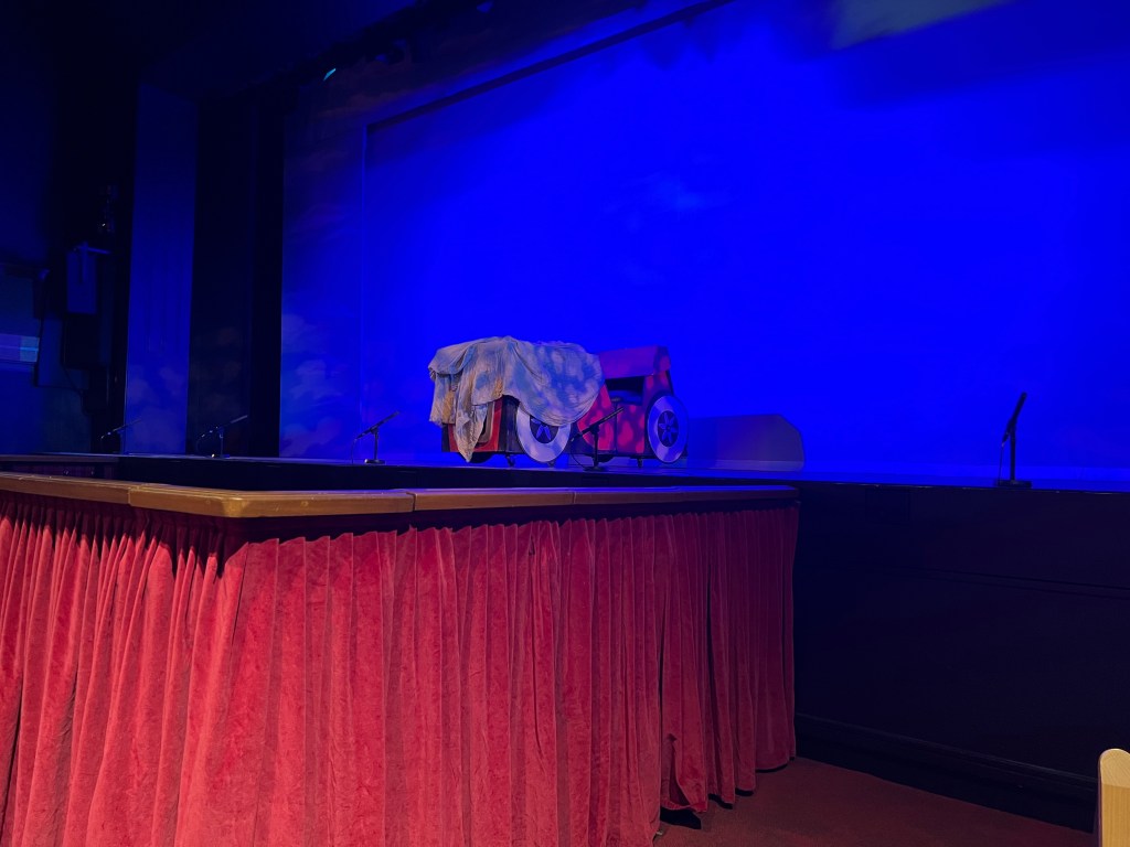 A picture of the stage before the show, with a bright blue background with an old fashioned red car cover in a old green sheet.