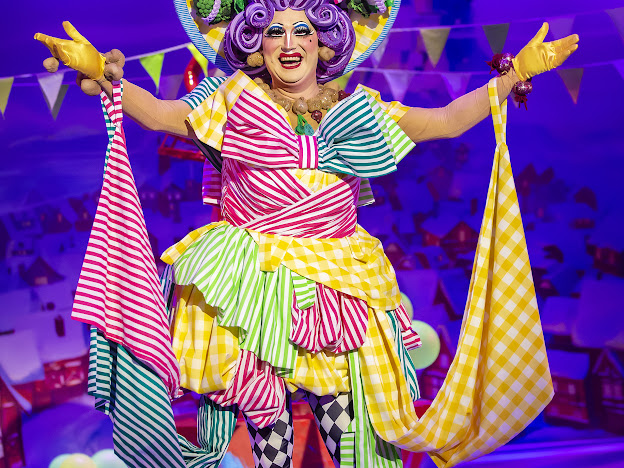 The picture shows a person dressed in a vibrant and colorful costume, seemingly performing on a stage. The person is wearing a dress made of different patterns including stripes and checks, in bright colors like pink, yellow, green, and blue. They have a big smile on their face and are wearing exaggerated makeup, including purple eyeshadow and bright red lipstick. They also have a curly purple wig that is adorned with green leaves and yellow fruits. They are extending their arms wide open and holding the ends of the dress, which is flowing. The background is blurry but seems to be a stage with a festive setting, including bunting flags.