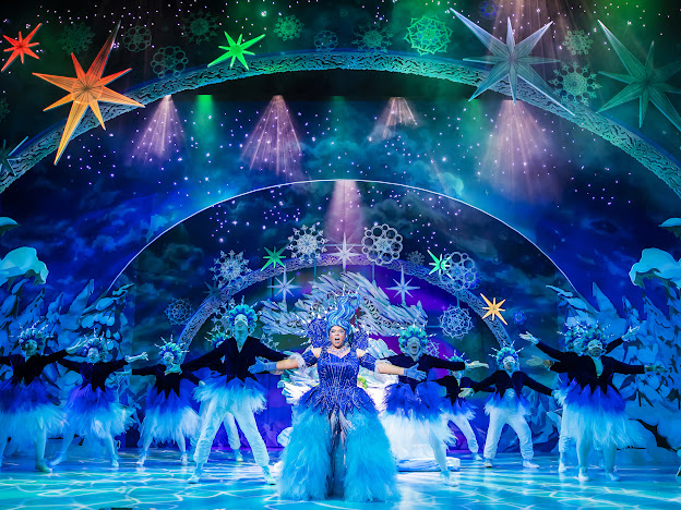 The picture is of a vibrant and colorful stage performance. The background is a magical night sky with stars and snowflakes, illuminated by beams of light in shades of purple and blue. There is a large arch on the stage adorned with snowflakes and stars.
In the center of the stage, there is a female performer wearing a sparkling blue dress with a voluminous skirt made of blue feathers. She has her arms outstretched and appears to be singing or performing.
Surrounding her are other performers dressed in black with blue feathered skirts. They are in various dance poses with their arms raised and appear to be part of a chorus or ensemble.
The overall feel of the image is festive, magical, and full of energy.
