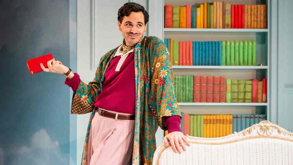 The image shows a man standing in a room with a playful and confident expression. He is holding a red book in his left hand and resting his right hand on the back of a white, ornate sofa. He is dressed in a colorful outfit: a teal robe with a floral pattern, a maroon sweater, pink pants, and a brown belt. Behind him is a bookshelf filled with neatly arranged books in various bright colors. The room has a light blue wall and a grayish-blue floor.