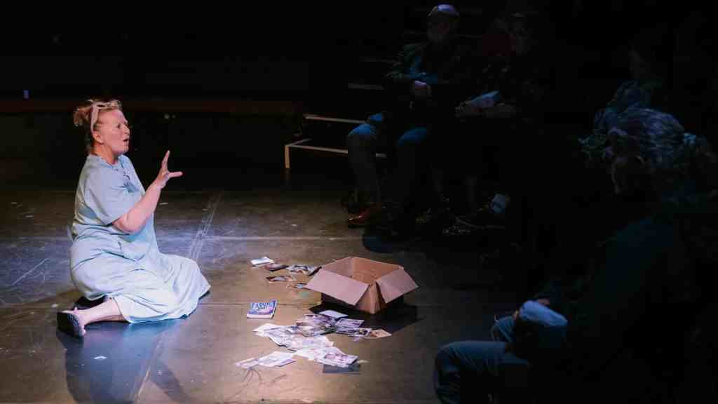The image shows a theatrical performance with a single actress on stage. She is sitting on the floor, wearing a light blue hospital gown, with her hair up in a messy bun. Her expression is animated, and she appears to be in the middle of delivering a dialogue or monologue. In front of her, there is an open cardboard box with scattered photographs all around it on the stage floor. The audience is visible in the background, sitting in darkness, attentively watching the performance. The setting seems intimate, with the audience close to the performer.