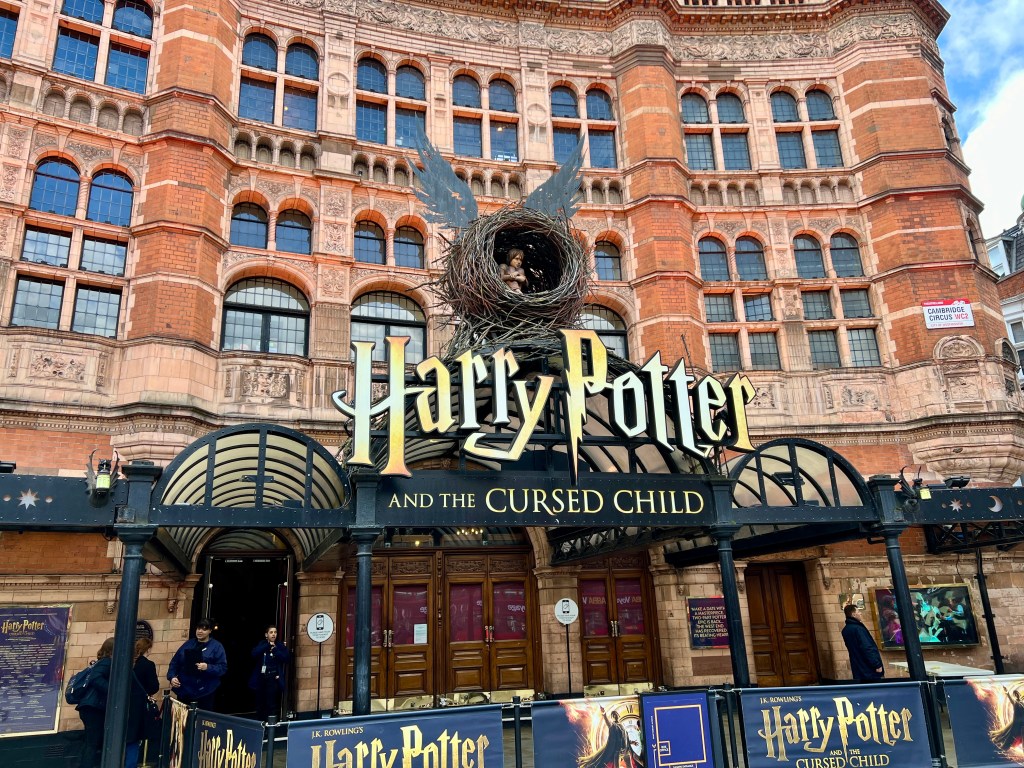 The picture shows the front facade of the Palace Theatre, currently hosting the play "Harry Potter and the Cursed Child". The theater has a classic Victorian red brick design with ornate stone carvings and arched windows. Above the entrance, there is a large, black sign with gold lettering that reads "Harry Potter" and beneath it, in smaller letters, "and the Cursed Child". Above the sign, there is a nest-like structure with a swirl that resembles the iconic 'snitch' from Harry Potter. Below the sign, there are two sets of double doors with a semi-circular glass canopy over the entrance. There are a few people around the entrance, and promotional banners for the play are visible on the railings in front of the theater.