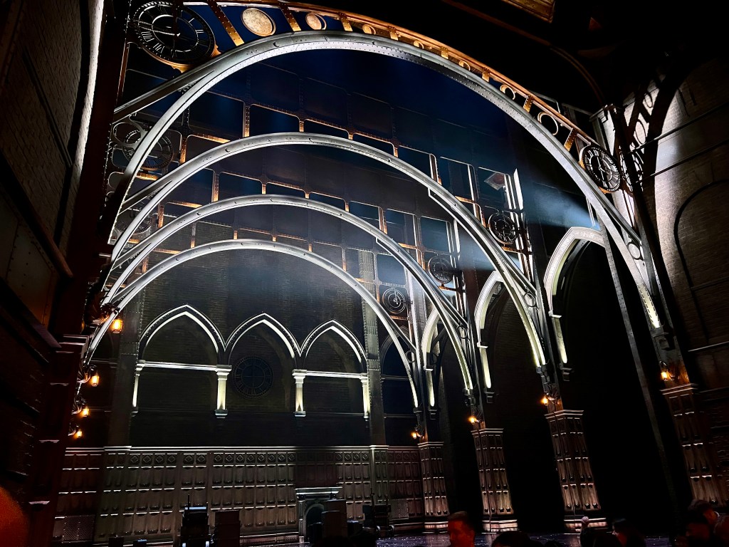 The picture shows an ornate interior with a gothic architectural style. There are a series of arches that ascend in size from the foreground to the background. The arches are made of metal with decorative elements and are illuminated by ambient lighting, which gives a dramatic and elegant effect. The walls are dark with patterns that resemble brickwork, and there are pointed arch windows with tracery. The overall atmosphere is grand and theatrical, possibly a setting for a performance or an event. There are some people seated at the bottom of the image, suggesting this is a public space.