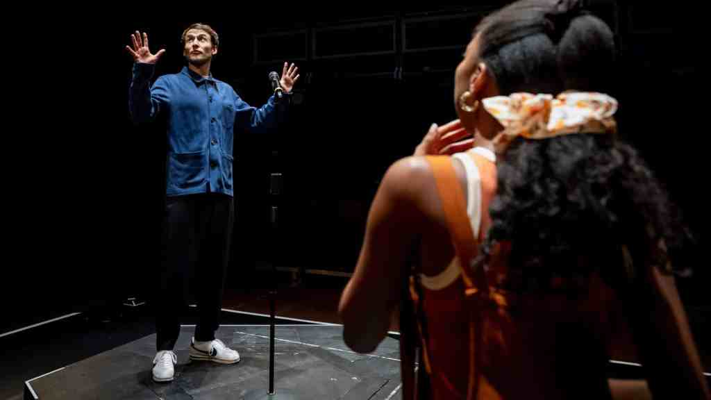 The image depicts an indoor scene with two individuals. A male figure stands in the background, wearing a dark blue jacket, with his hands in his pockets. He stands in a relaxed posture, looking towards the foreground. In the foreground, there is a female figure seen from the back. She has dark hair, which is tied back with a large bow accessory, and is wearing an orange sleeveless top. The scene appears to be dimly lit, suggesting an intimate or dramatic setting, possibly in a theater or a similar performance space. The overall mood seems contemplative and focused, indicating a moment of interaction or confrontation between the two characters. The background is dark, with indistinct details, but it reveals hints of red seats, further reinforcing the impression of a theater environment.