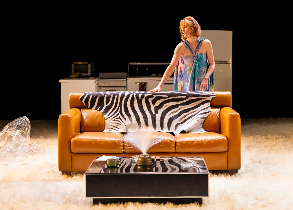 The image depicts an indoor scene with a woman standing behind a tan leather sofa, which is covered with a zebra-print blanket. She is wearing a colorful, flowing dress and appears to be adjusting the blanket. In front of the sofa, there is a coffee table with a gold-colored ornament that looks like a fountain, spraying water upwards. Behind the woman, there is a kitchen setup with a white refrigerator, a microwave, and a stove. The floor is covered with a fluffy, white carpet, and the background is dark, suggesting the image might be part of a performance or artistic setup.