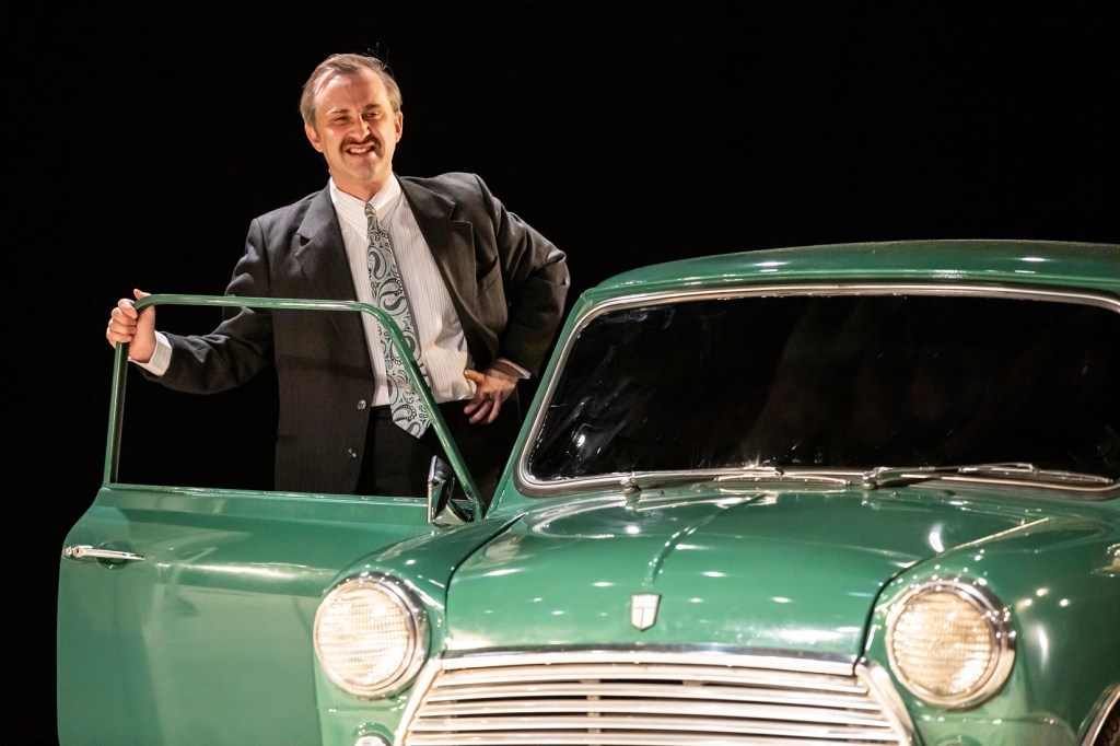 The image shows a man standing next to a vintage green car with its driver's door open. He is dressed in a dark suit and tie, and his right hand is resting on his hip while his left hand holds the car door. The background is dark, emphasizing the car's shiny green exterior and the man's formal attire. The car's headlights and curves suggest it is a classic model, adding a nostalgic and elegant touch to the scene. The overall atmosphere is one of sophistication and timeless style.