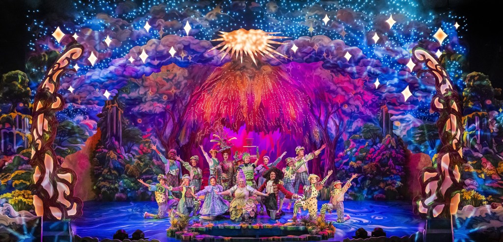 This image depicts a vibrant stage performance with a fantastical theme. Centered on the stage, a group of performers, including men and women dressed in elaborate costumes with floral and nature-inspired designs, are frozen in a celebratory pose with their arms raised. The background features a dramatic and colorful scenery of an enchanted forest, illuminated with bright, shimmering lights resembling stars, and a radiant burst of light or fireworks at the top center, adding a magical atmosphere. The mood conveyed is joyous and festive, enhanced by the rich colors and dynamic arrangement of the performers.