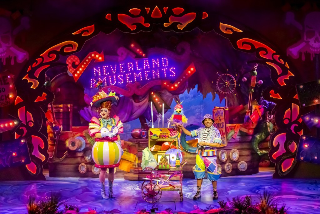 The image depicts a vibrant, colorful stage scene from a circus or amusement-themed performance titled 'Neverland Amusements.' In the center, under a bright purple and pink sign, stand two performers dressed in elaborate, whimsical costumes. The performer on the left, possibly female, wears a yellow, red, and green striped outfit with a puffy skirt, while the performer on the right, possibly male, is in a multicolored outfit with blue pants. They are surrounded by a variety of props, including a decorated cart filled with playful objects. The background is adorned with bright lights, creating an atmosphere of excitement and wonder.
