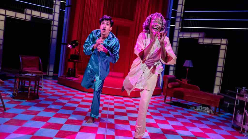 Two actors stand on a brightly lit stage with a red curtain backdrop. The floor has a red and blue checkerboard pattern. Both performers are mid-dance, smiling and animated. The person on the left wears shiny blue silk pyjamas, while the person on the right wears a light pink robe and trousers. Behind them are vintage-style furniture pieces, including a record player, a red armchair, and a lamp.