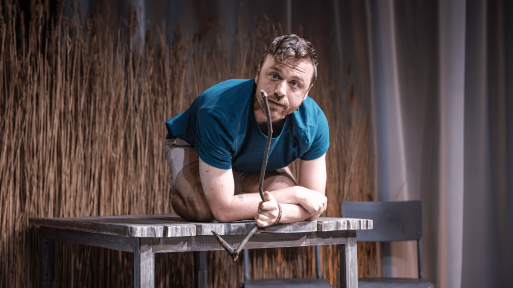 A man crouches on top of the same wooden table, holding a long stick in his hands and leaning slightly forward as if pretending to draw in the sand or poke at something on the ground. He has short hair and a thoughtful, focused expression, with a hint of curiosity or playfulness. He’s wearing a short-sleeved blue T-shirt and beige shorts. Behind him, the same tall golden reeds create a natural, rustic backdrop, softly lit in warm tones. The lighting feels gentle and earthy, suggesting an outdoor or beach-like setting.