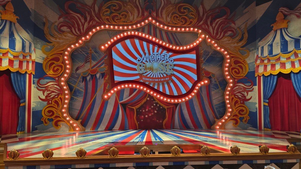 Image shows a inside of a Circus tent pantomime set with a giant screen the middle showing the title mother goose! The colours are the tradition circus colours of red white and blue