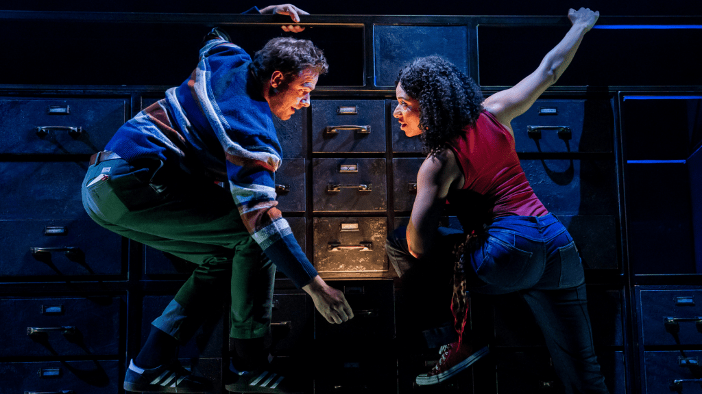 The image depicts an indoor scene with two individuals, one male and one female, interacting with a large metal filing cabinet structure that spans the background. The female, positioned on the right, has curly black hair and wears a sleeveless red top with tight dark jeans and sneakers. She is climbing the cabinet, holding onto it with one hand while looking intently in the direction of the male. The male, on the left, is dressed in dark pants, sneakers, and a striped jacket and is similarly interacting with the cabinet, appearing to be in a mid-climbing posture. His focus is directed downward toward an open drawer illuminated by a warm light emitting from inside. The filing cabinet itself is comprised of multiple small, square drawers in a matte black color, creating a geometric pattern. The overall environment is dimly lit, with focused lighting highlighting the characters and the intrigue of the open drawer, suggesting a mysterious or intense mood. The activity appears dynamic, with the characters engaged in an exploration or search, giving the scene a sense of urgency or curiosity. The atmosphere conveys suspense and collaboration, heightened by the interplay of shadows and light.