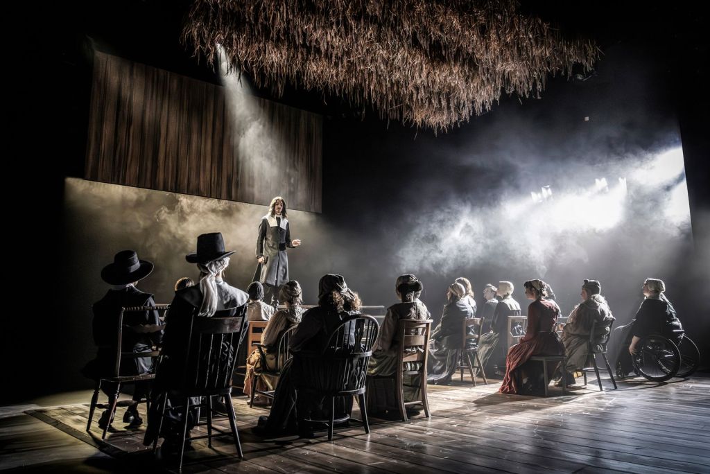 A theater stage scene with a group of people seated in chairs facing a standing speaker under dramatic lighting and fog. The setting looks like a period play: many people wear old-fashioned clothing such as bonnets, shawls, long dresses, and wide-brimmed hats. The speaker stands slightly elevated near the center-left, wearing a long dark coat and light shirt, with long hair, as if addressing the seated group.

A strong spotlight shines down from above onto the speaker, and additional bright stage lights from the right create heavy haze and smoke across the scene. The floor is wooden planks, and the audience on stage sits in several rows of mismatched wooden chairs. A large textured element hangs overhead, resembling a thatched or straw ceiling piece, adding to the rustic atmosphere.