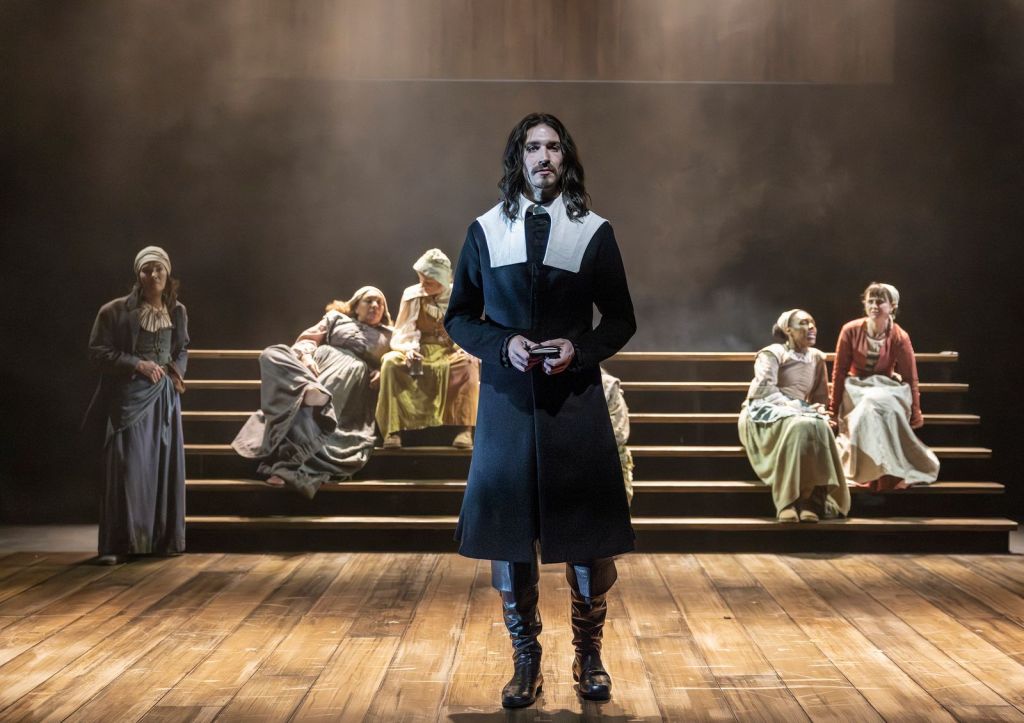 A theater stage scene with a man standing in the center foreground under warm spotlighting, and several women seated on tiered wooden steps behind him. The man has light skin and long dark wavy hair, and wears a long dark coat with a wide white collar and tall dark boots. He is facing forward with a serious expression and holding a small object in both hands at waist level. Behind him, women in muted, old-fashioned dresses and head coverings sit or lean on the steps, spaced across the background. The stage floor is wooden, and the back of the stage is dark with haze or smoke catching the light.