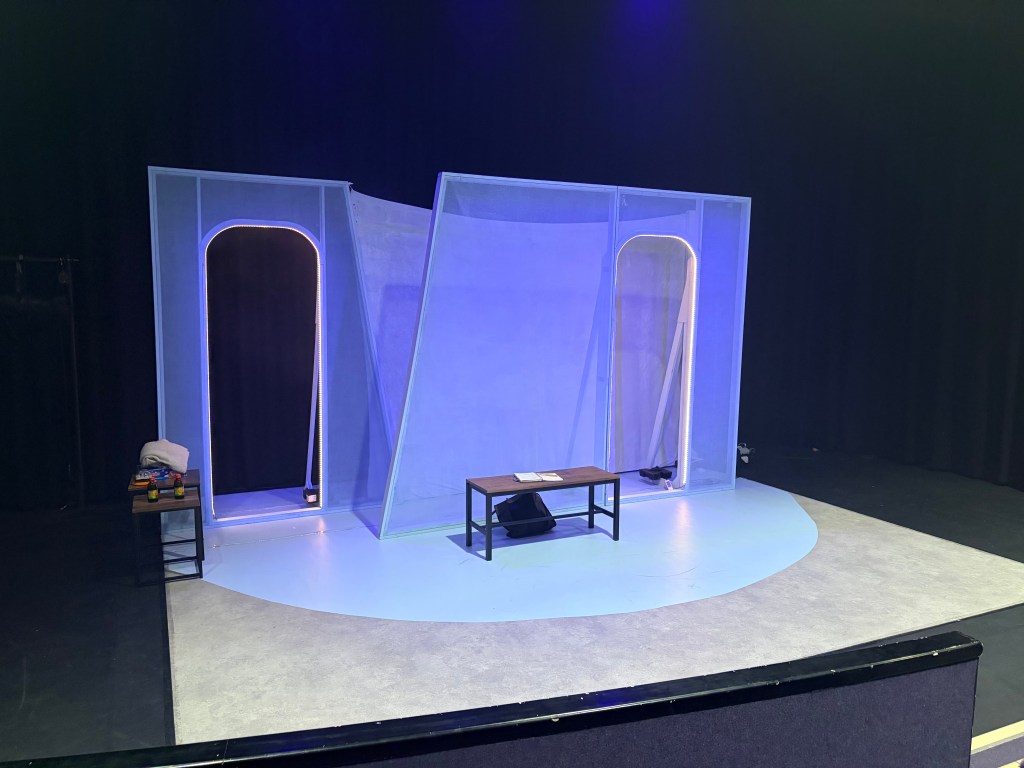 A minimalist theater stage set with cool blue lighting.  
There are three large, slightly angled translucent wall panels across the back of the stage; the left and right panels each have a tall rounded-rectangle doorway outlined with vertical LED strip lights that glow white with a hint of warm color. The center panel is solid and leans slightly, giving the whole wall a stylized, off-kilter look.  

On the floor in front of the wall is a pale blue oval or rounded shape that contrasts with the surrounding light gray stage floor. At the center of this blue area stands a simple rectangular wooden coffee table with dark legs; on top of it are a couple of sheets of white paper. Under the table, near the middle, is a black object that looks like a small bag or case.  

To the far left, just in front of the wall, is a small side table with a dark metal frame and wooden top. On this side table are a folded light-colored towel or cloth and some colorful rectangular items, possibly books or props, along with two small bottles or containers.  

The background beyond the set is all black curtains, and the lighting on the set is primarily blue from above, giving the panels and floor a soft, cool glow while the surrounding stage remains darker.