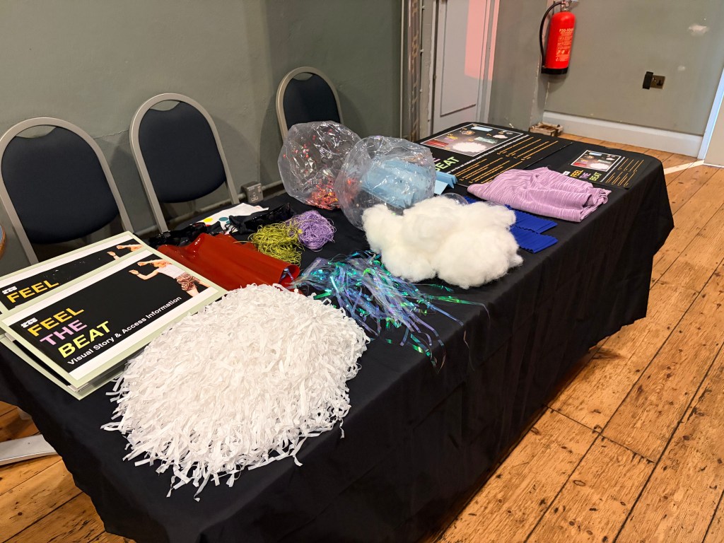 A rectangular table covered with a black cloth filled with sensory and promotional items.  
On the left side of the table, two large display boards lie flat; they are mostly black with yellow and purple text that reads “FEEL THE BEAT” and “Visual Story & Access Information,” and they each have a photo of a person moving to music.  
In front of these boards is a large, mound-like mass of shredded white plastic or paper strips, like a big pom‑pom.  
Next to it are several sheets of shiny red and orange plastic material laid out flat.  
Further along the table, there are tangles of thin cords or strings in neon yellow and purple colors.  
In the middle of the table sits a clear plastic bag partially filled with small, multi‑colored, reflective pieces, giving a sparkly effect, and beside it there is another clear bag containing light blue soft material.  
Near the center‑right is a fluffy pile of white stuffing or cotton‑like material, with some loose, iridescent fiber‑optic style strands and blue and green ribbon‑like strands spread beneath or around it.  
To the right are several rectangular blue textured pads stacked together, and above them is a folded piece of pale purple, finely striped fabric.  
Three grey plastic chairs with dark seats are lined up behind the table.  
In the background on the right, a red fire extinguisher is mounted on the wall, and the floor is wooden with visible planks.