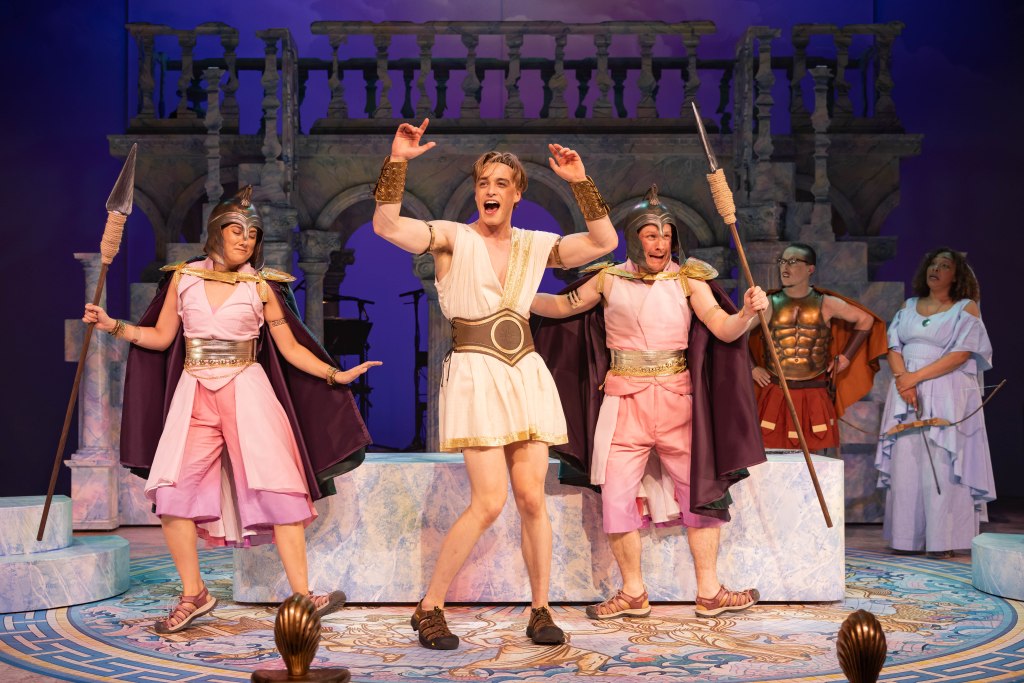 A colorful stage musical scene with actors dressed in ancient Greek-style costumes.  
In the center, a young light-skinned man with short blond hair stands on a round marble-patterned platform. He wears a white sleeveless tunic with a deep V-neck, gold trim, and a wide brown and gold belt. His arms are raised enthusiastically, mouth open in mid-song or shout, and he wears brown sandals and gold wrist bracers.  

On his left and right are two light-skinned performers in matching costumes: pink sleeveless jumpsuits with split skirts, gold belts, metallic gold shoulder pieces, dark purple capes, and bronze-style helmets with crests. Each holds a tall spear upright. The performer on the left poses with one arm extended and knees bent in a dance stance. The one on the right has an animated expression, mouth open, with the spear angled slightly forward.  

Behind them, slightly to the right, stands a person with short dark hair, glasses, and medium skin tone wearing a bronze muscle armor chest plate, red knee-length skirt panels, and an orange cape, watching the central trio. Further right is a woman with medium-brown skin and curly shoulder-length hair, dressed in a light blue, flowing, off-the-shoulder gown with layered ruffles. A bow is in her hand and a quiver of arrows hangs at her side.  

The set is designed like a marble Greek temple, with pale blue and pink marbled columns, arches, and a balcony rail across the back. The floor has an ornate circular design in blues and golds with stylized patterns. The background lighting is purple, giving the scene a fantastical, mythological atmosphere.