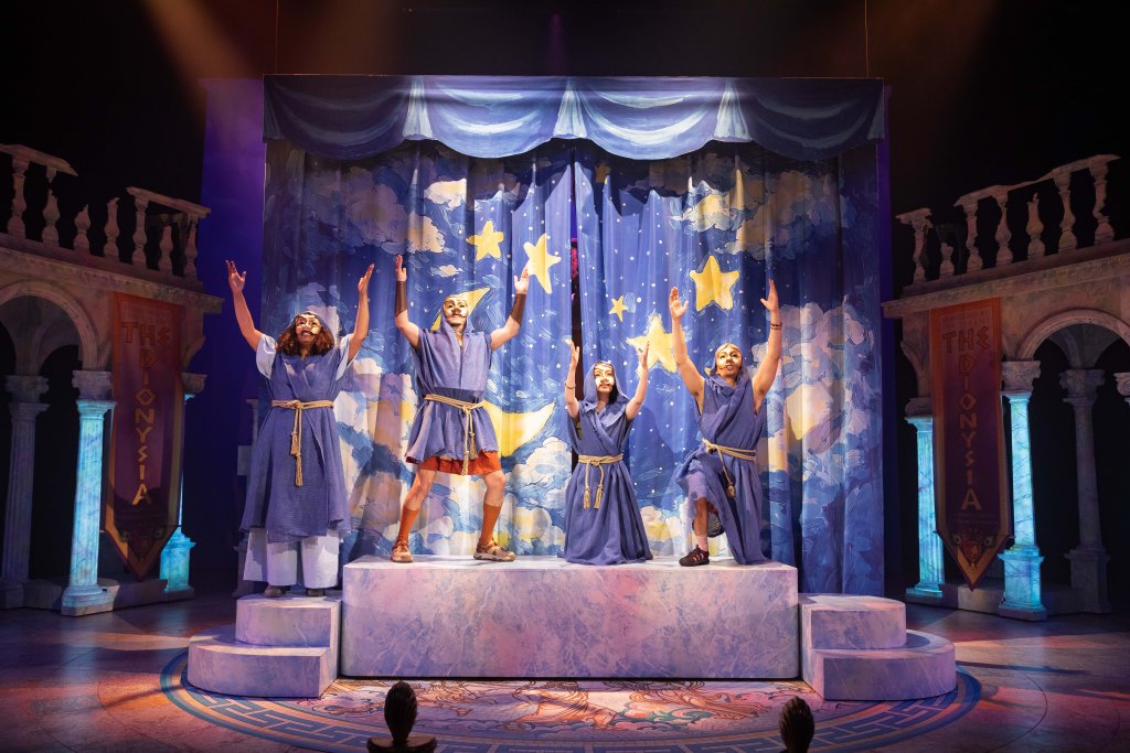 Colorful stage performance with four actors standing on marble-like platforms.  
They are in mid-pose with arms raised, as if celebrating or singing.  
All four wear matching blue tunic-style costumes tied with light-colored rope belts, and theatrical half-masks with exaggerated noses and expressions, reminiscent of ancient Greek comedy masks.  
The center actor’s tunic is shorter, showing bare legs with sandals; the others wear longer, robe-length tunics.  
Behind them is a large curtain painted with a night sky: deep blue background, clouds, yellow stars, and a crescent moon framed by draped blue “fabric” along the top.  
On both sides of the stage are cream-colored stone-like columns and arches suggesting an ancient Greek temple or theater.  
Tall vertical banners hang on each side, reddish-orange with the words “THE DIONYSIA” written vertically.  
Warm stage lighting from above creates a golden glow on the actors and platforms, with some purple and blue light on the backdrop.  
At the very bottom edge, the tops of a few audience members’ heads are just visible, facing the stage.