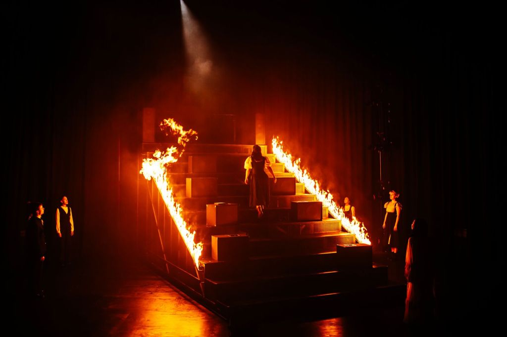 A large, dramatic staircase on a theater stage with both sides lined in tall orange flames.
The stairs form a wide pyramid shape, made of dark rectangular blocks that step upward to a flat platform at the top.
A single bright spotlight from above shines down toward the top center of the structure, cutting through haze or light smoke and emphasizing the heat of the fire.
On the staircase, a person in a dark dress with short sleeves is walking upward toward the top, facing away.
The flames are continuous along the outer edges of the stairs, rising several inches to a foot high, with bright yellow and orange tongues and darker smoke near the top.
The stage floor and background are very dark, almost entirely black, so the fire and the figures are the main visible elements.
On the lower left side of the image, two people stand on the stage floor, facing the staircase; both wear dark clothing, one in a suit-style outfit with a lighter shirt.
On the right side of the stage, two more people stand spaced apart, also facing the staircase; they wear sleeveless or short-sleeved dark outfits, their faces and arms lit by the orange glow.
In the front right corner, nearer to the camera, a figure in a long, light, semi-transparent garment stands watching the scene, mostly in shadow except for some orange highlights from the fire.
The overall atmosphere is intense and theatrical, with strong contrasts between the bright flames and the surrounding darkness, giving a sense of a climactic or ritual-like moment in a performance.