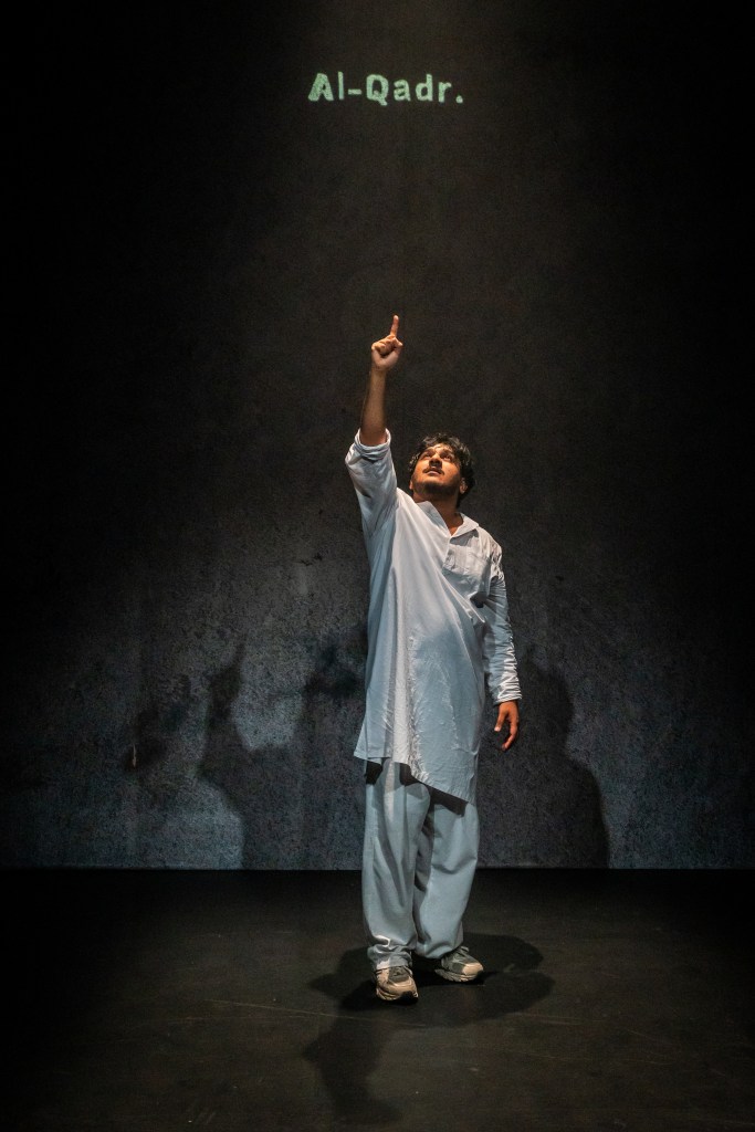 A man stands alone on a dimly lit stage, wearing loose light-blue traditional clothing and grey sneakers.
He is positioned slightly to the right of center, facing forward with his right arm raised straight up and his index finger pointing toward the ceiling.
His expression appears intent and focused, with his head tilted slightly back in the direction he is pointing.
Soft light from above highlights his face and upper body, casting a tall shadow behind him on the textured dark-grey wall.
Near the top of the image, projected text glows in pale greenish-white and reads: “Al-Qadr.”