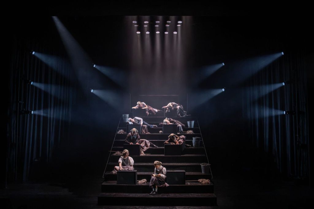 A stepped wooden platform on a dark stage with several performers arranged across its levels.
The platform rises like a wide staircase into the background, and each level holds one or two performers dressed in muted brown and gray clothing that suggests historical peasant or servant attire, including long skirts, aprons, and headscarves.
Most performers are slumped, lying, or sitting in weary poses, some leaning on boxes placed on each step as if they are workstations or beds.
Metal buckets and piles of what looks like rags or mop heads sit beside some of the boxes, hinting at cleaning or labor.
Bright, cool-toned stage lights shine diagonally from both sides toward the center and down from above, cutting through haze so that the light beams are clearly visible and framing the performers in a dramatic, somber atmosphere.
The surrounding stage area beyond the lit platform is mostly in darkness, with only faint outlines of curtains and side lighting rigs visible, so attention is focused on the central group.